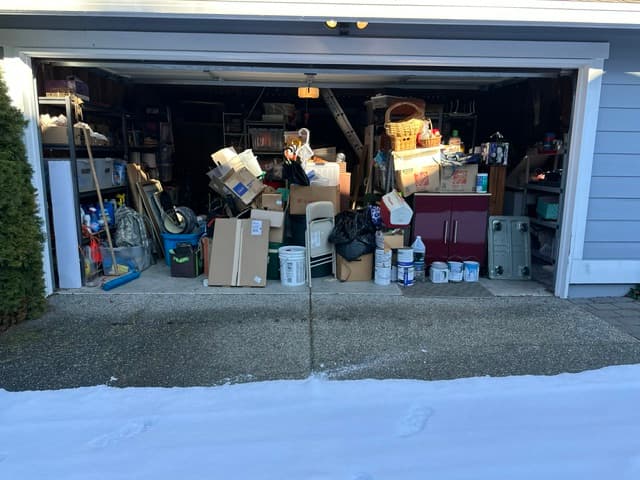A cluttered garage packed with boxes, paint cans, and household items behind a snowy driveway.