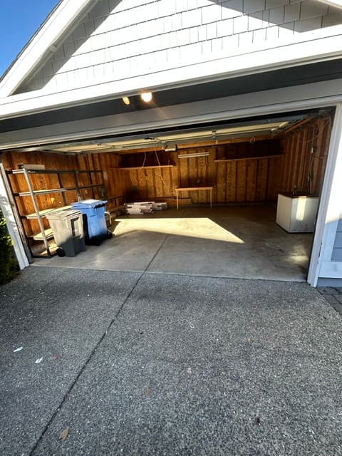 Open garage with wooden walls, metal shelving, trash bins, and sunlight on concrete floor.