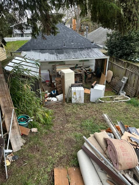 Cluttered backyard with old appliances, construction debris, and building materials near a small shed.