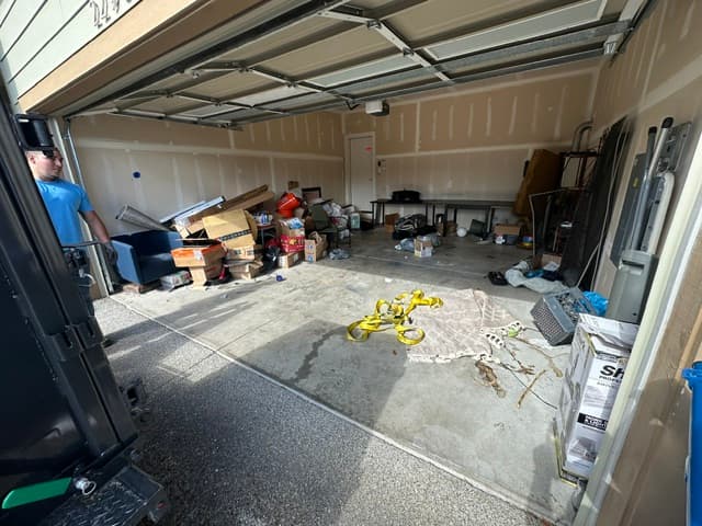 A man stands beside a cluttered garage filled with cardboard boxes, furniture, and yellow straps.