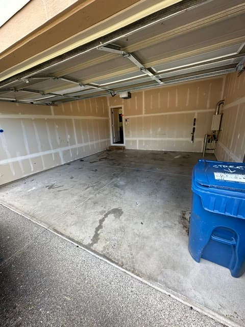 Empty garage featuring unfinished drywall, concrete floor, blue recycling bin, and an open doorway.