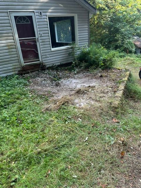 Gray house with white door and window beside a cleared dirt patch in overgrown grass.