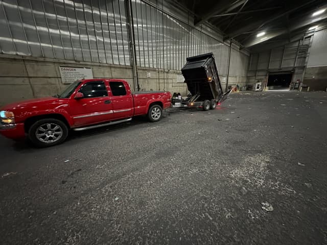 Red pickup truck towing a black dump trailer with its bed raised in a warehouse.